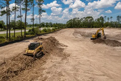 Excavator clearing a multi-acre commercial site with graded sand in the foreground and a treeline awaiting removal beyond