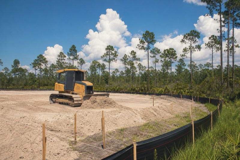 Bulldozer rough-grading a cleared sandy lot with silt fence staked along the property edge under a summer sky