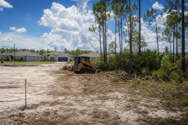 Freshly cleared quarter-acre residential lot with sandy soil exposed, wooded uncleared lots visible on both sides